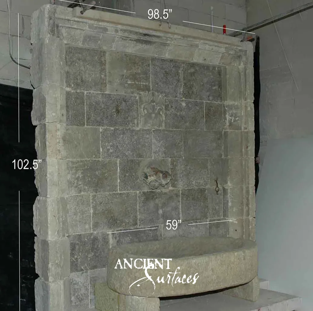 Wide reclaimed antique limestone wall fountain from Ancient Surfaces photographed in the warehouse, a grey ashlar backplate between pale projecting side piers with a carved palmette, a worn mascaron spout and a semicircular basin on stone supports