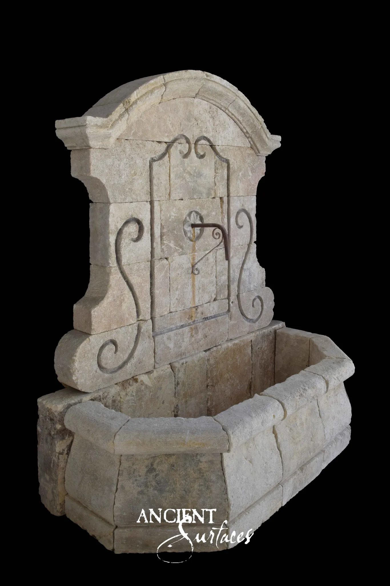 Cream reclaimed antique limestone wall fountain from Ancient Surfaces with a segmental arched cap, long S scrolled volute wings down each side, an applied iron scrollwork panel with a small spout and a long canted corner trough