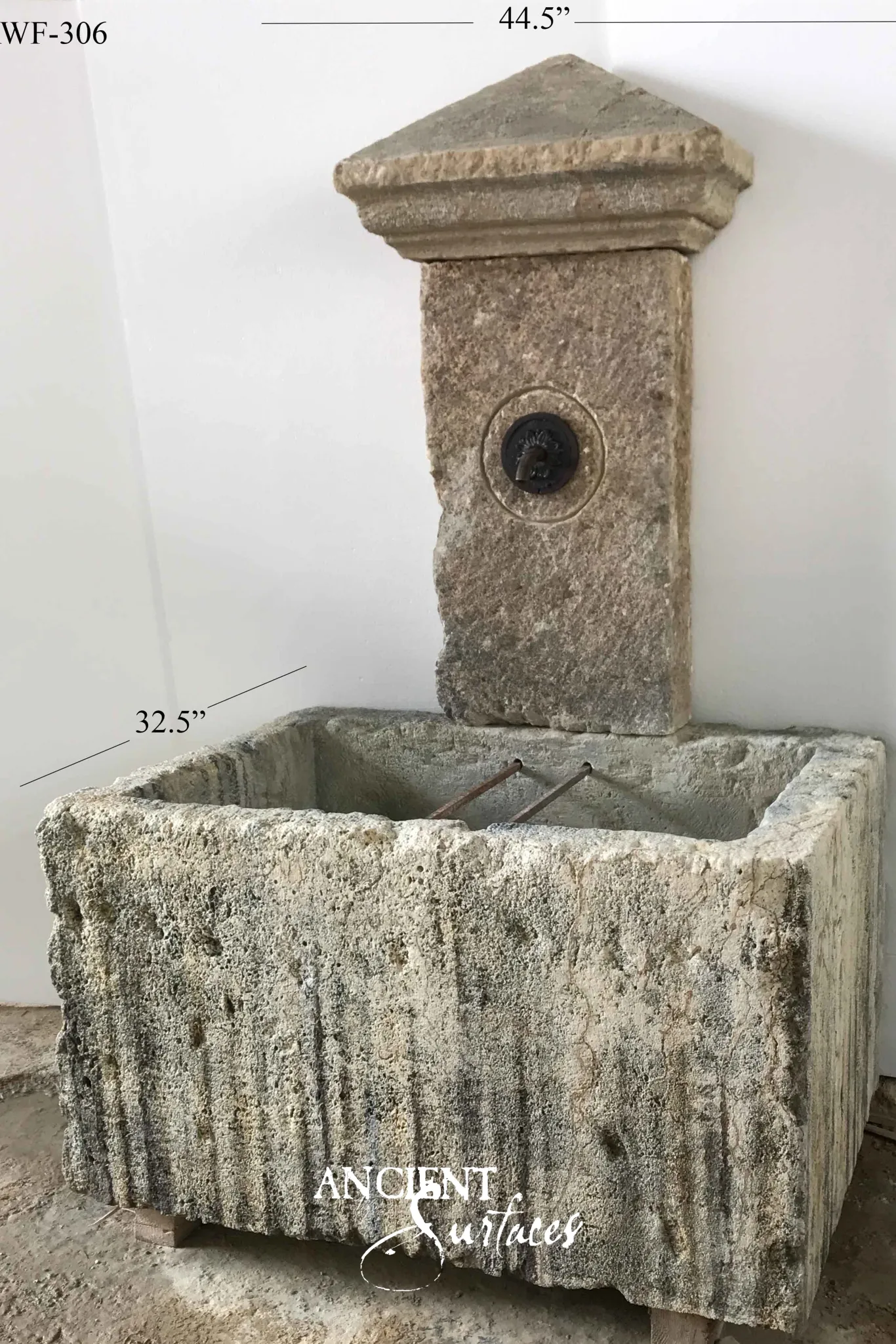 Narrow reclaimed antique limestone wall fountain from Ancient Surfaces, a pier with a triangular pediment cap and a bronze lion boss spout set in a round recess, above a monolithic trough scoured into deep vertical runnels