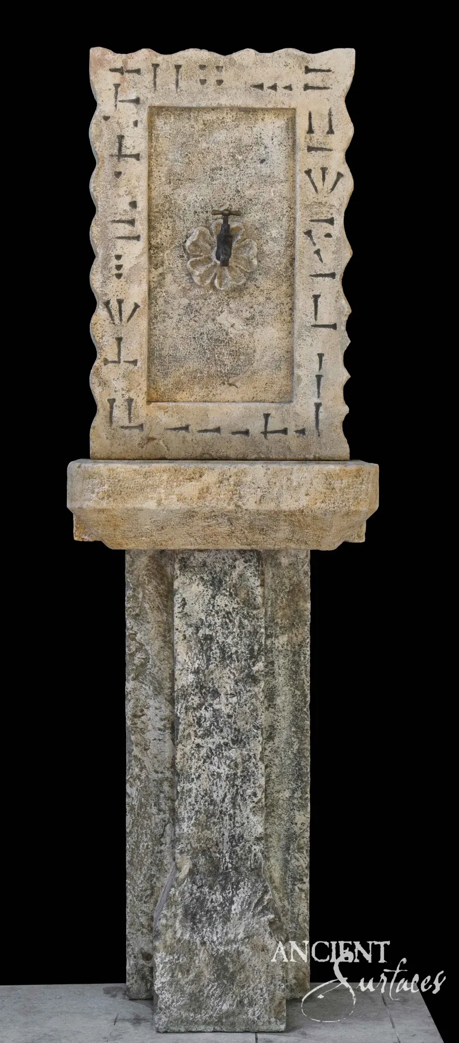 Tall reclaimed antique limestone wall fountain from Ancient Surfaces with a scalloped border chiselled with repeating incised bracket and hatch marks, a carved rosette boss and bronze spout, on a shallow basin carried by a rough limestone pier