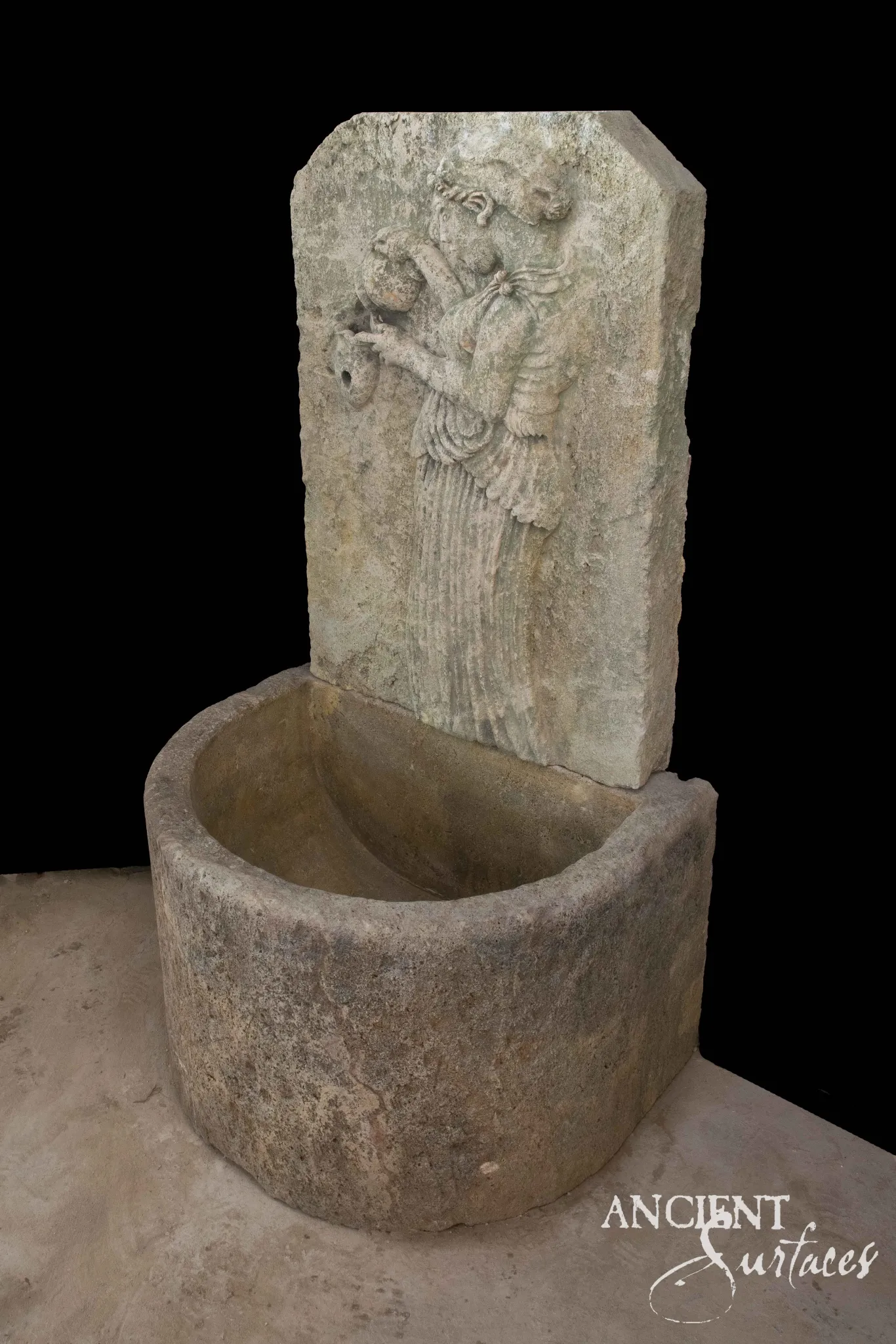 Reclaimed antique limestone wall fountain from Ancient Surfaces, a single relief carved slab showing a robed figure pouring from a vessel in deep drapery folds, above a heavy semicircular basin cut from one block