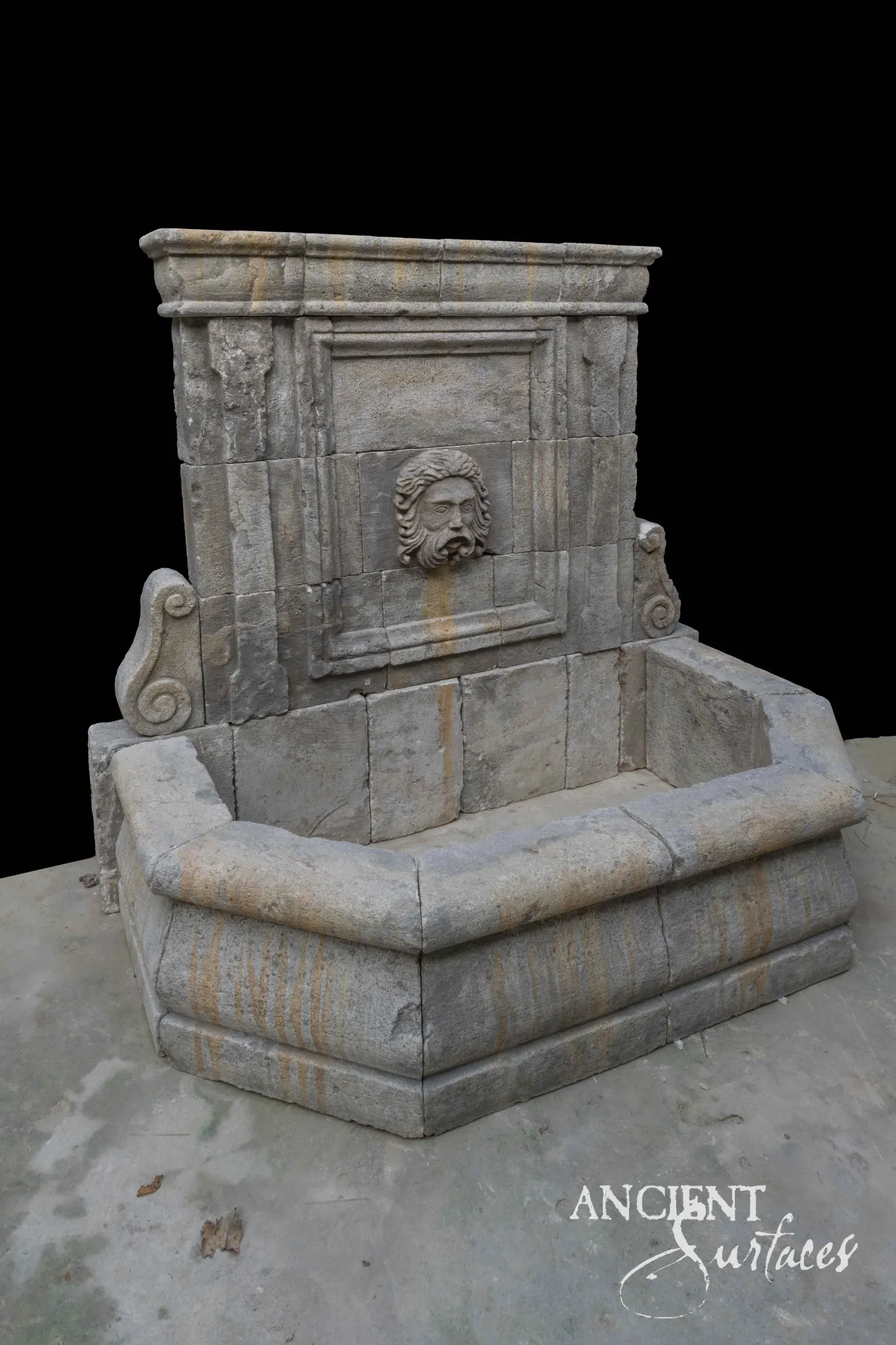 Reclaimed antique limestone wall fountain from Ancient Surfaces with a bearded mascaron spout carved in high relief, set in a moulded fielded panel between scrolled volute consoles above a wide canted basin banded with ochre veining