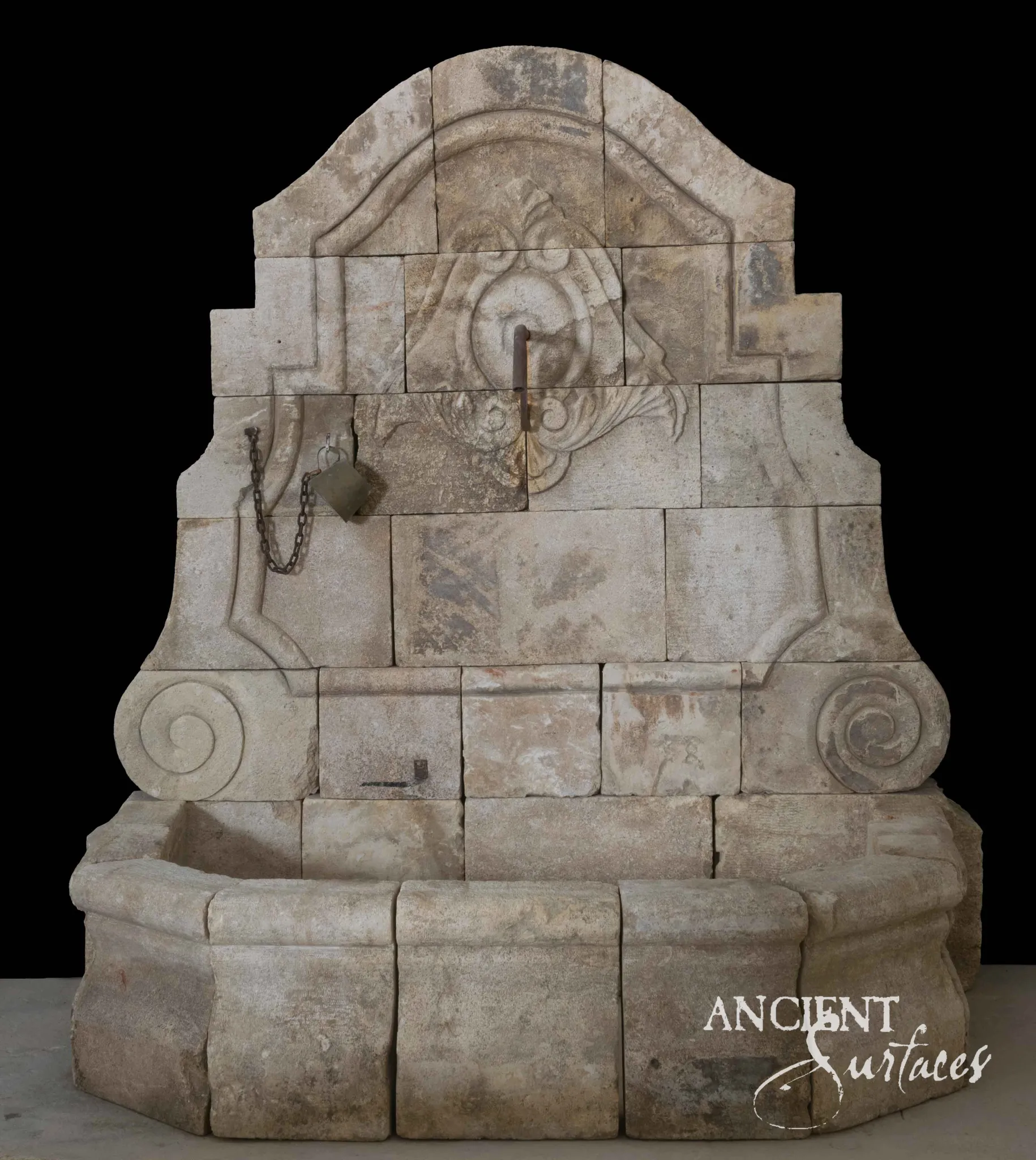 Tall pale reclaimed antique limestone wall fountain from Ancient Surfaces with a shell and acanthus cartouche in the arched crest, large spiral volutes, a hanging iron chain and counterweight and a broad canted apron basin