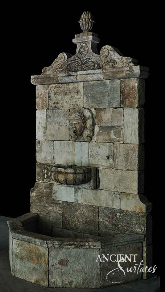 Tall reclaimed antique limestone wall fountain from Ancient Surfaces crowned by a carved artichoke finial over a broken scrolled pediment, a heavily mottled ashlar backplate, a lion mask spout and a small gadrooned half bowl
