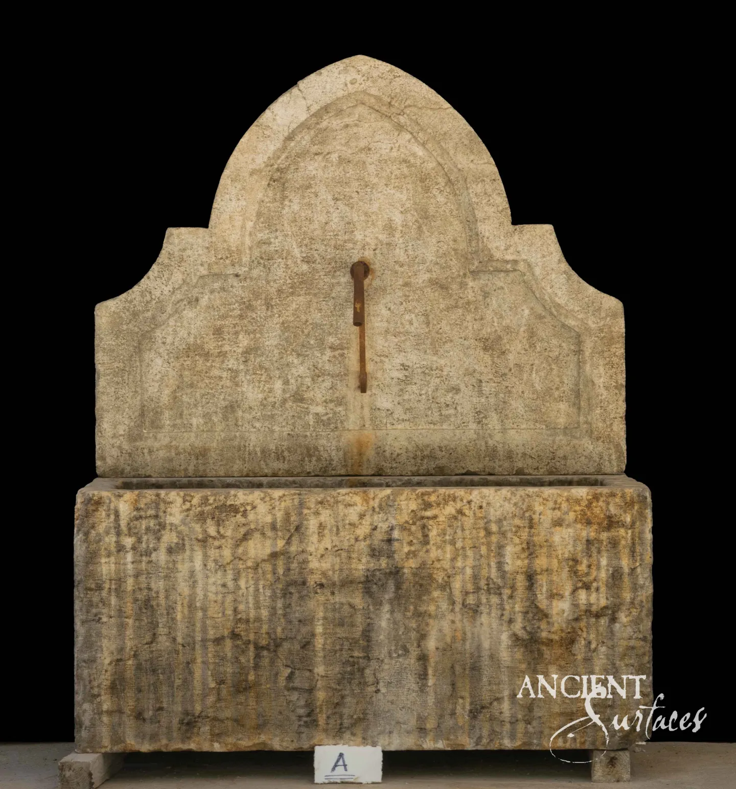 Undecorated reclaimed antique limestone wall fountain from Ancient Surfaces, a single slab cut to an ogee arch with stepped shoulders and a short iron spout, above an enormous monolithic trough scoured into vertical striations