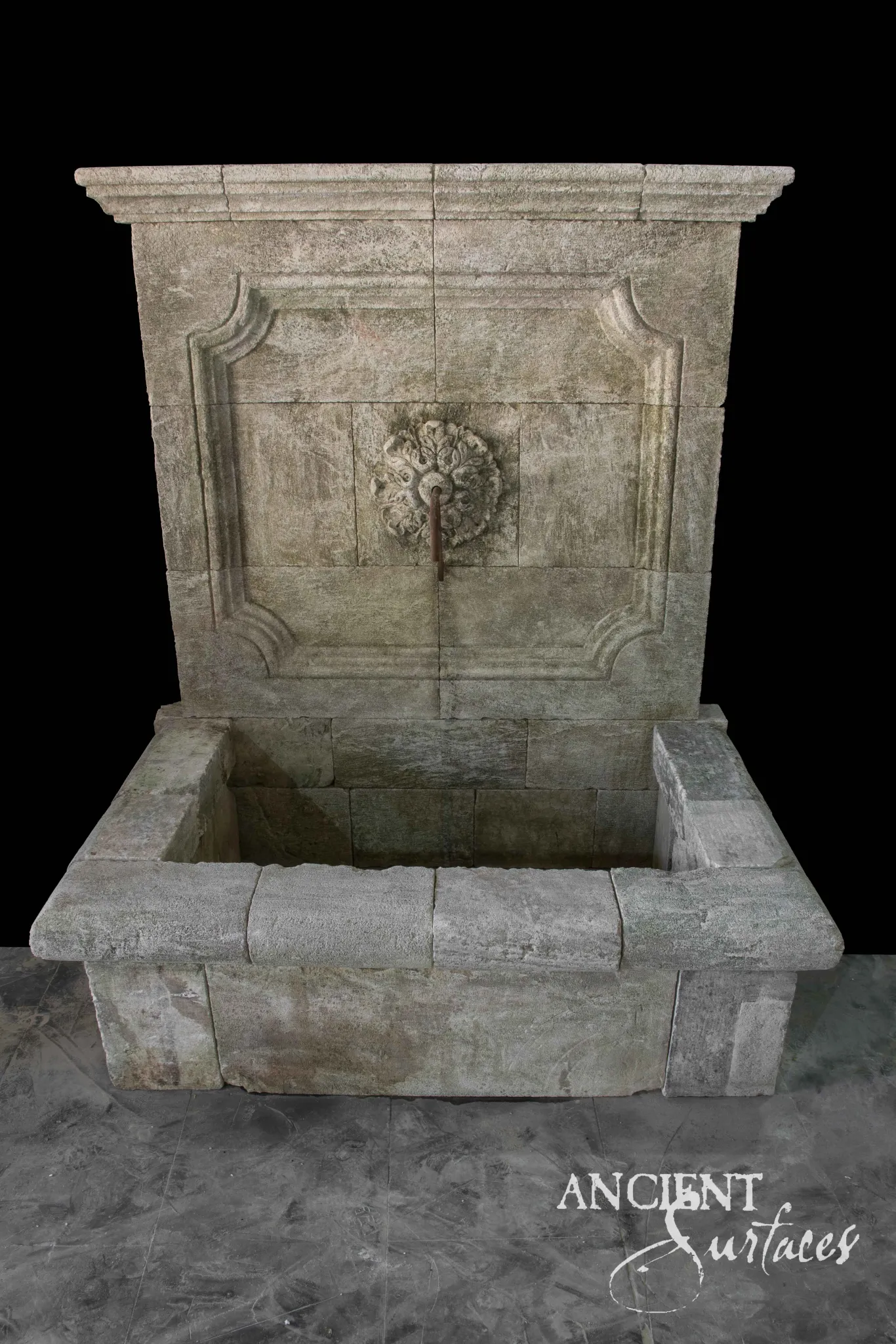 Reclaimed antique limestone wall fountain from Ancient Surfaces in soft grey green stone, a deep cornice over a recessed panel with cusped scrolled corner brackets, a carved foliate rosette spout and a block built basin with moulded coping