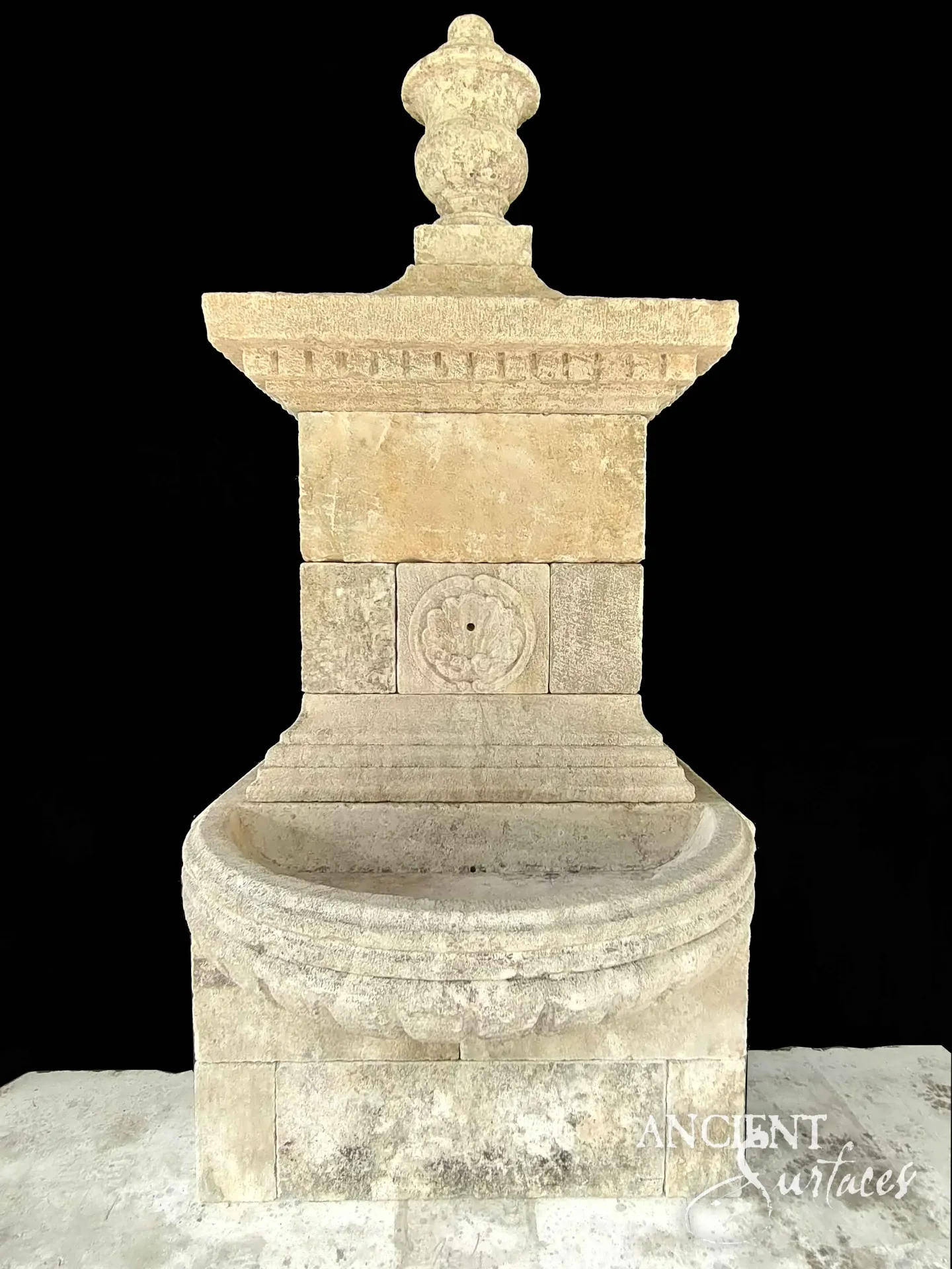 Honey gold reclaimed antique limestone wall fountain from Ancient Surfaces crowned by a turned baluster finial over a dentil carved cornice, with a square rosette boss holding the spout and a semicircular gadrooned bowl below