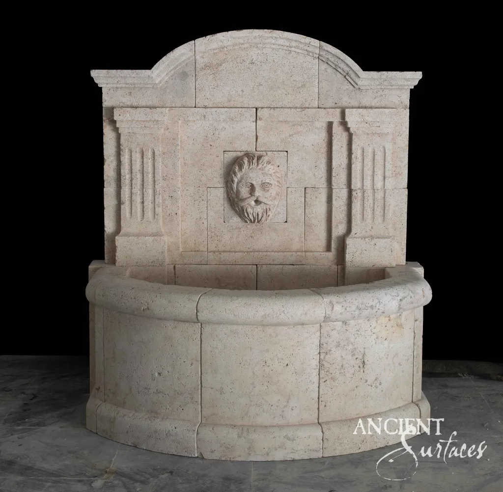 Pale finely pitted reclaimed antique limestone wall fountain from Ancient Surfaces with fluted pilaster strips framing a stepped cruciform panel, a maned leonine mask spout and a bowed semicircular basin with a rolled coping