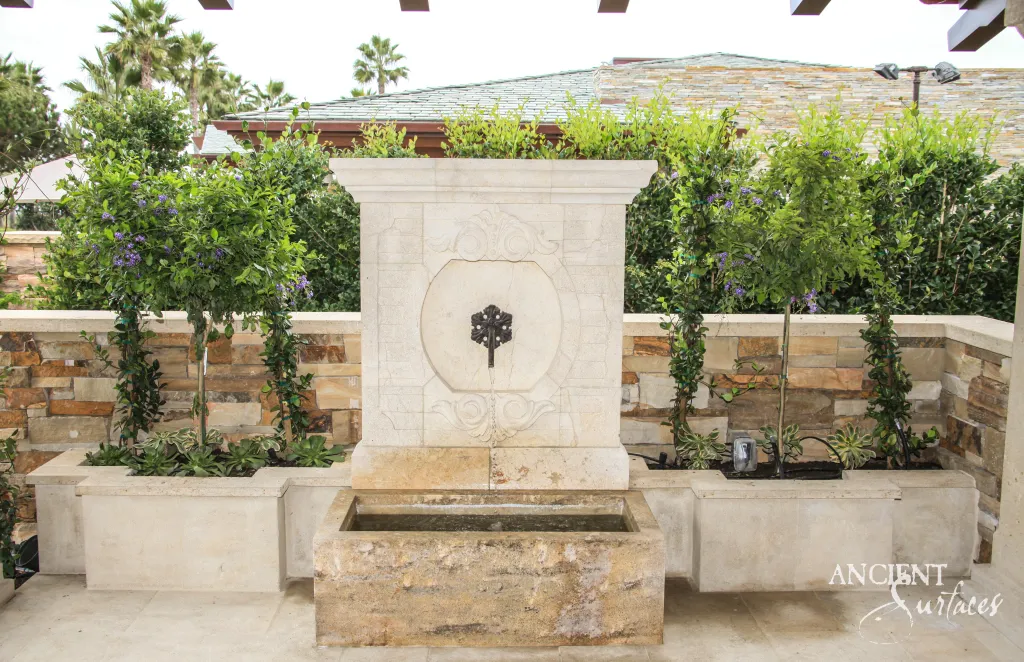 Reclaimed antique limestone wall fountain from Ancient Surfaces running on a terrace, its backplate carved with a large oval cartouche and foliate scrollwork around an ornate bronze cruciform spout, above a gold monolithic trough full of water