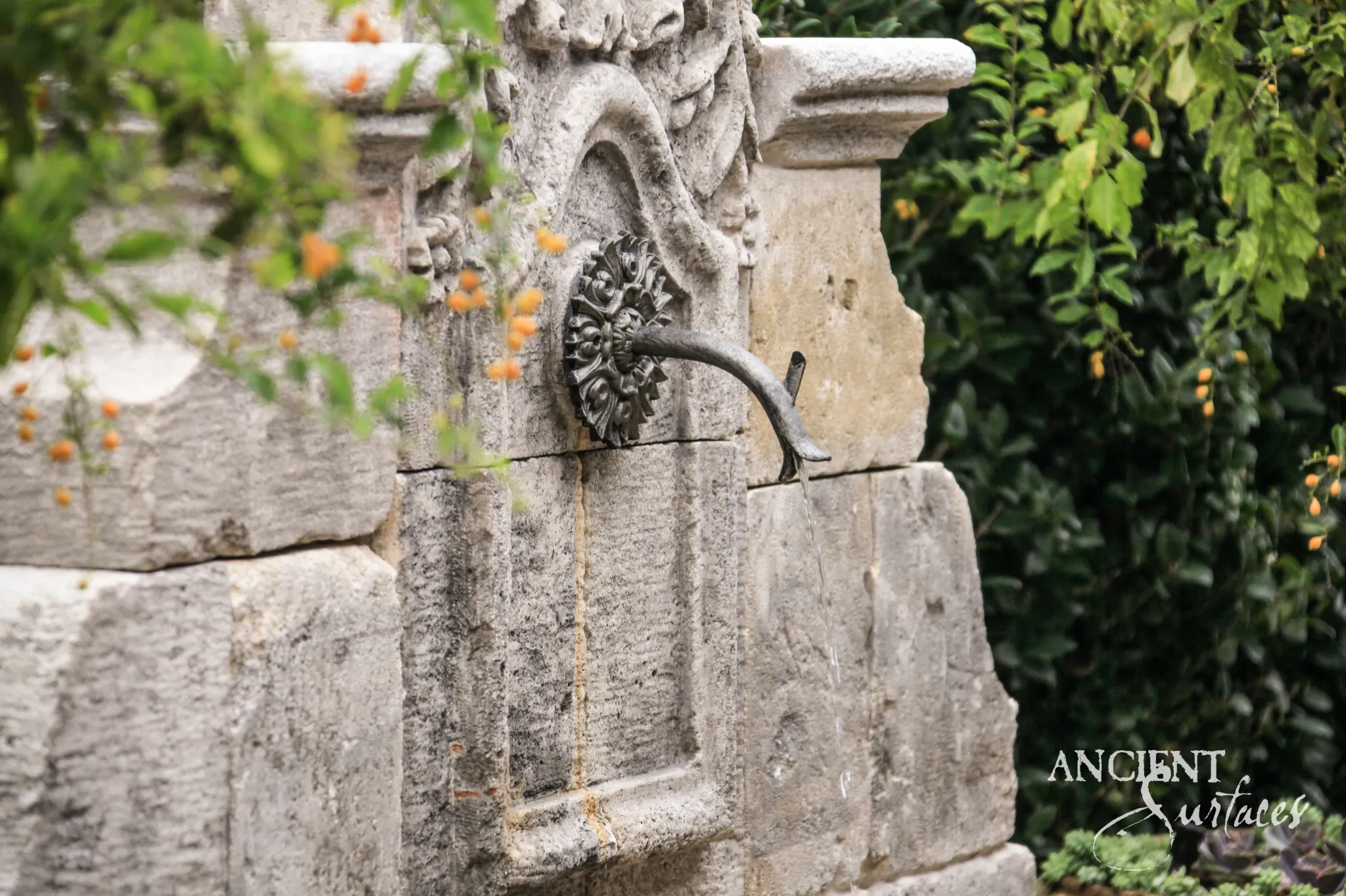 Reclaimed antique limestone wall fountain from Ancient Surfaces seen close, a cast bronze rosette escutcheon and curved gooseneck spout fixed to deeply pitted pale limestone streaked with rust runnels beneath a carved scrolled crest