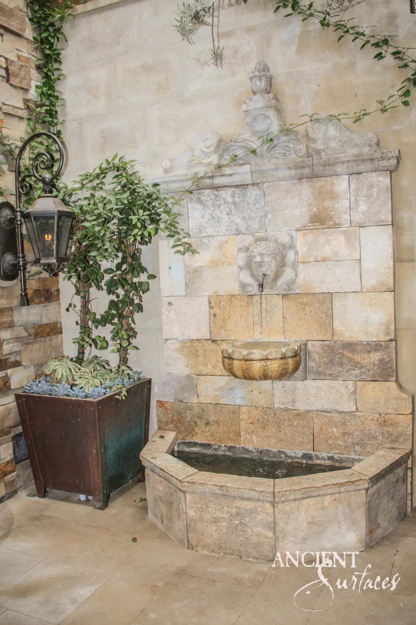 Reclaimed antique limestone wall fountain from Ancient Surfaces running in a plastered courtyard, water falling from a carved lion mask into a gadrooned half bowl and a canted apron basin beneath a broken pediment and artichoke finial